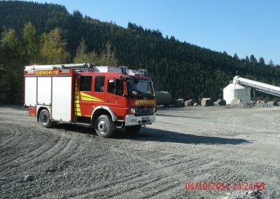 Ein Feuerwehr LKW steht in einen Steinbruch , im Hintergrund ein riesen Wald der dicht mit bäumen bewachsen ist, rechts im Hintergrund ein Silo mit förderband für Schutt beim Schadenpräventionstraining