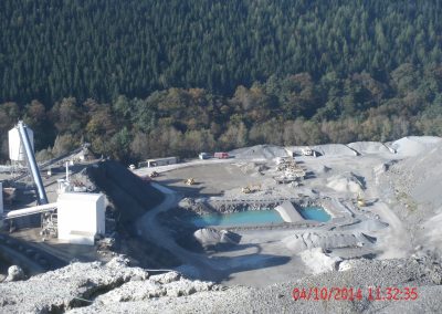 Ganzer Steinbruch vom höchste Punkt aus fotografiert, In der Mitte eine Quelle mit türkis scheinenden Wasser, links davon förder Bänder und Silos und Rechts hohe Steinruch Wände und im Hintergrund ein dicht bewachsener Wald