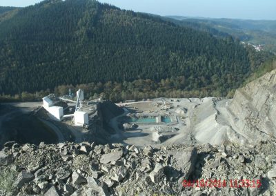 Ganzer Steinbruch vom höchste Punkt aus fotografiert, In der Mitte eine Quelle mit türkis scheinenden Wasser, links davon förder Bänder und Silos und Rechts hohe Steinruch Wände und im Hintergrund ein dicht bewachsener Wald